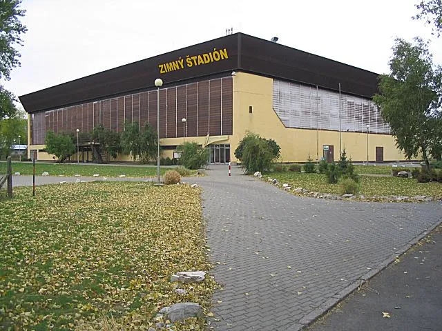 Zimný štadión Levice zvonku: dlhá hnedo-krémová hala s nápisom ZIMNÝ ŠTADIÓN na okraji strechy a dláždeným prístupom pred budovou. – Zimný štadión Levice