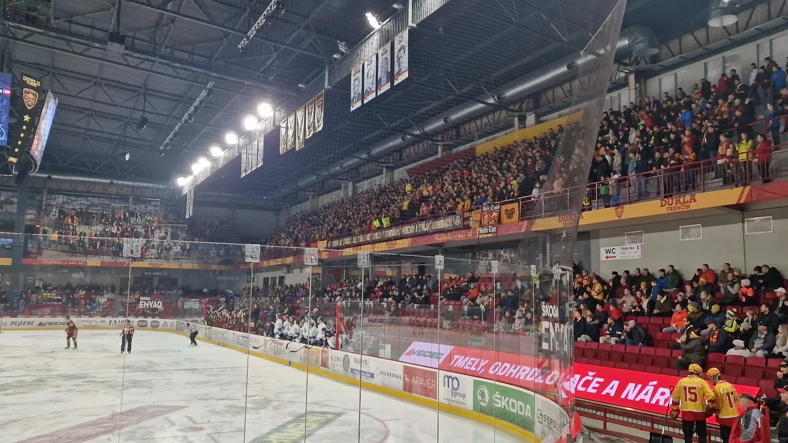 A Dukla Trenčín game under the scoreboard in a packed Zimný štadión Pavla Demitru, seen from the glass at ice level — Zimný štadión Pavla Demitru