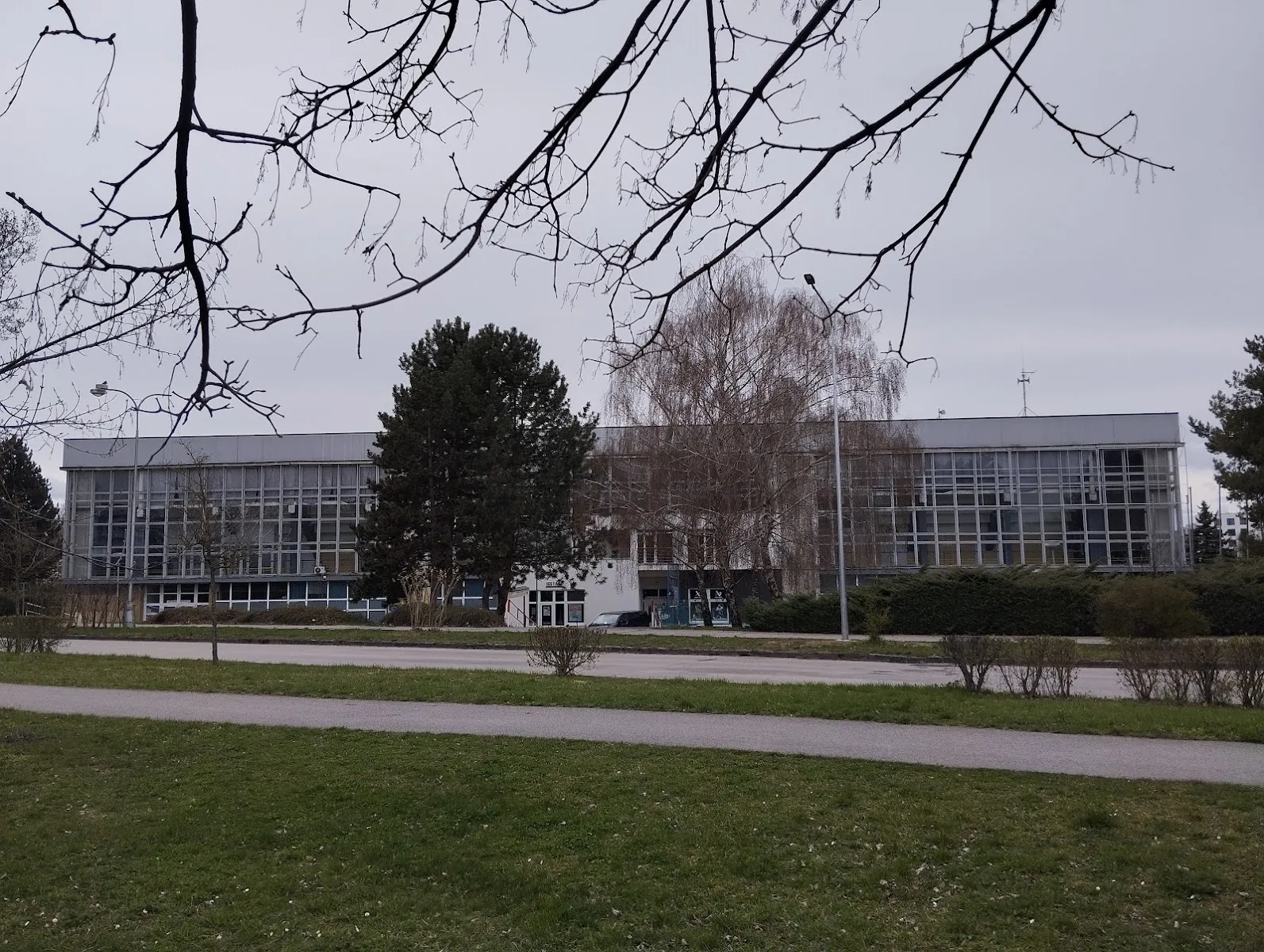 The low modern building of the Piešťany winter stadium seen across a lawn with bare trees. – Zimný štadión Piešťany