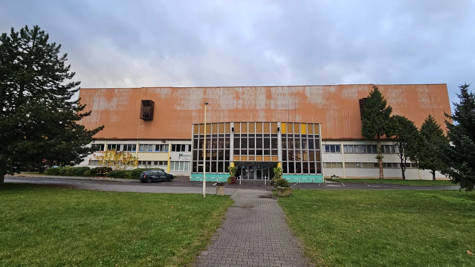The long low Prievidza winter stadium with an orange roof and a paved approach across a lawn. — Zimný štadión Prievidza