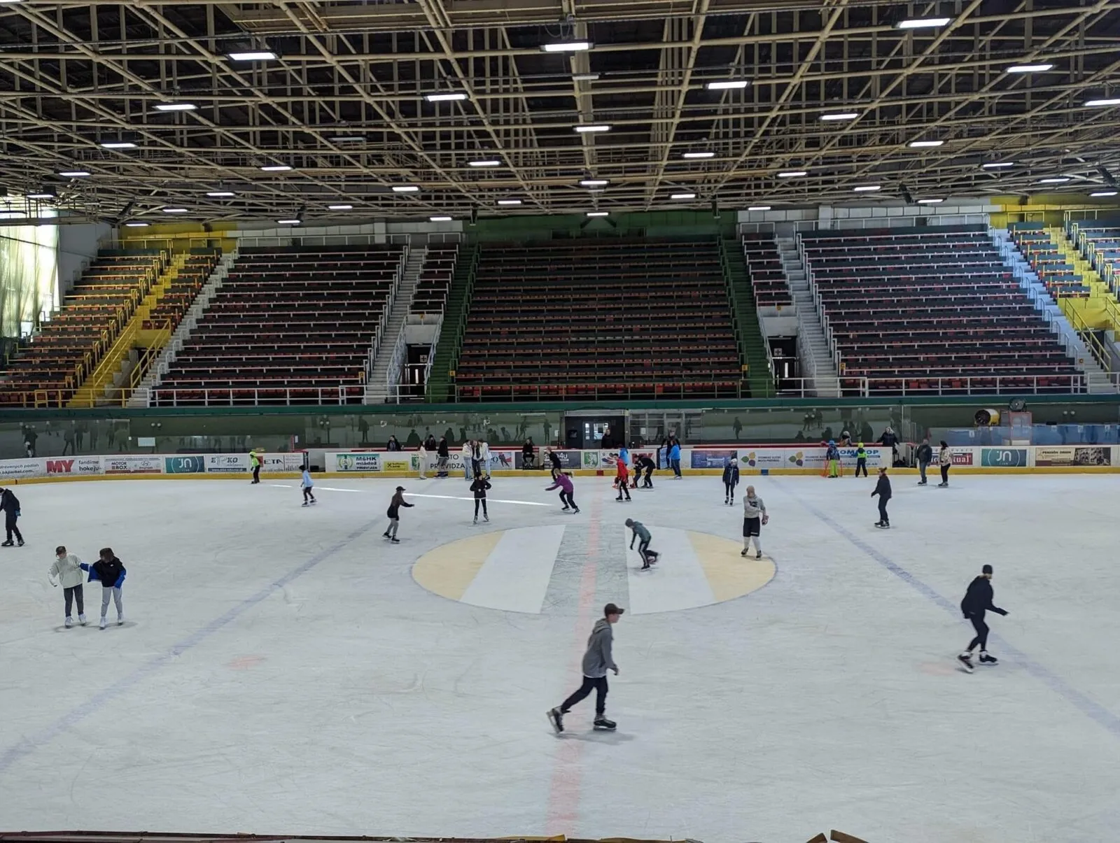 The ice sheet of the Prievidza winter stadium during public skating, with skaters spread across the ice and the empty stands and roof trusses behind. — Zimný štadión Prievidza