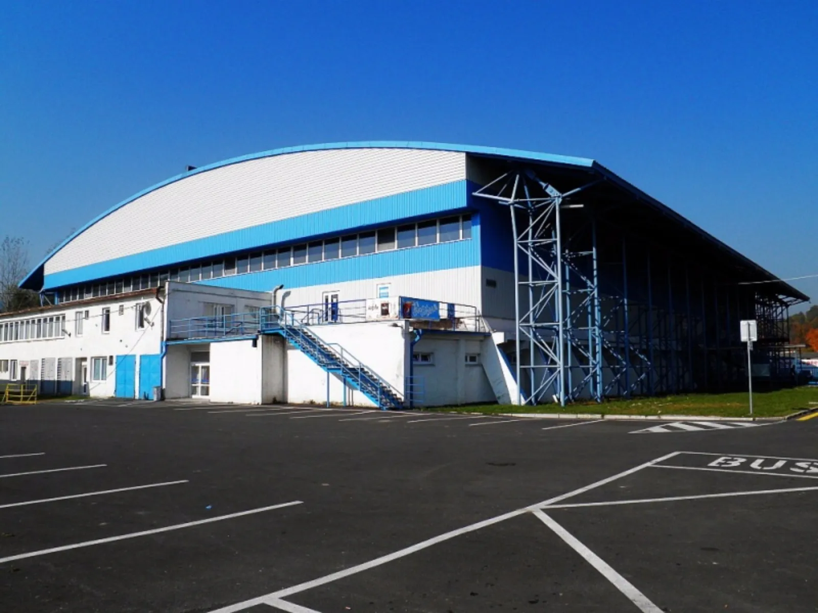 The curved white and blue roof of the Púchov winter stadium seen from the car park on a sunny day, with the stand structure along its side. — Zimný štadión Púchov