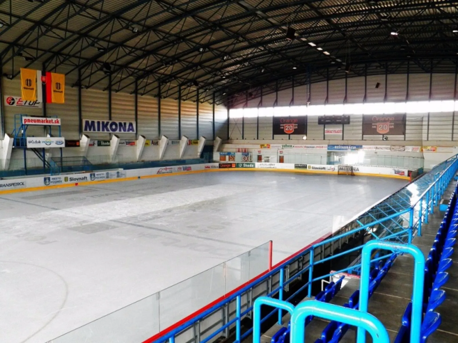The empty ice sheet of the Púchov winter stadium seen from the stand, with the glass-topped boards and blue seats along the side. — Zimný štadión Púchov