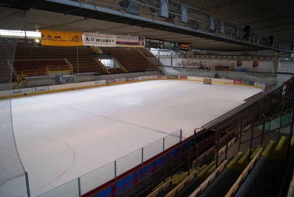 The empty ice of Zimný štadión Senica seen from the stands, with the covered seating along the far side and the roof trusses overhead — Zimný štadión Senica