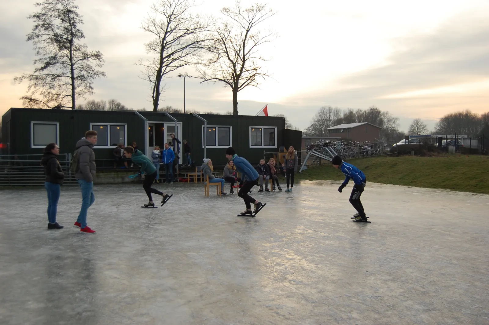 Schaatsers op afstand op het grijze natuurijs van de baan in Zwammerdam bij schemering, met de donkergroene clubhuishut en kale bomen aan de overkant. – IJsbaan Zwammerdam (Zwammerdamsche IJsclub)